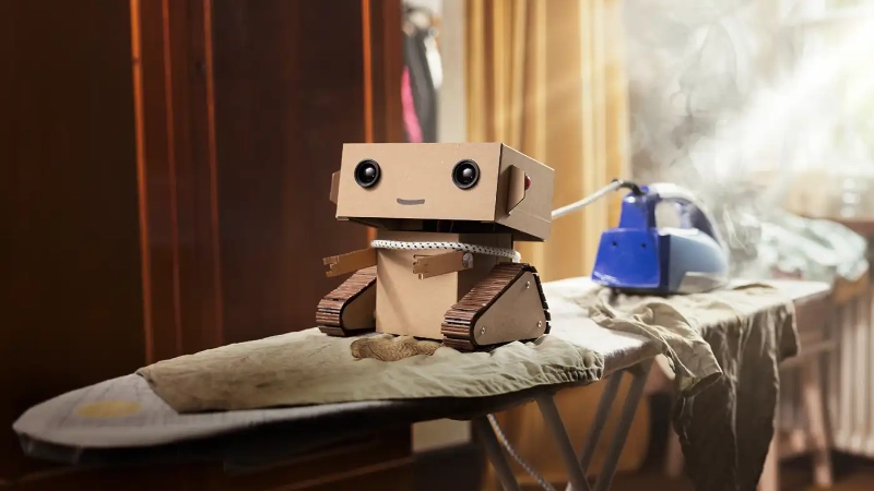 Der Kartonroboter Robi versucht, Kleidungsstücke zu bügeln.
