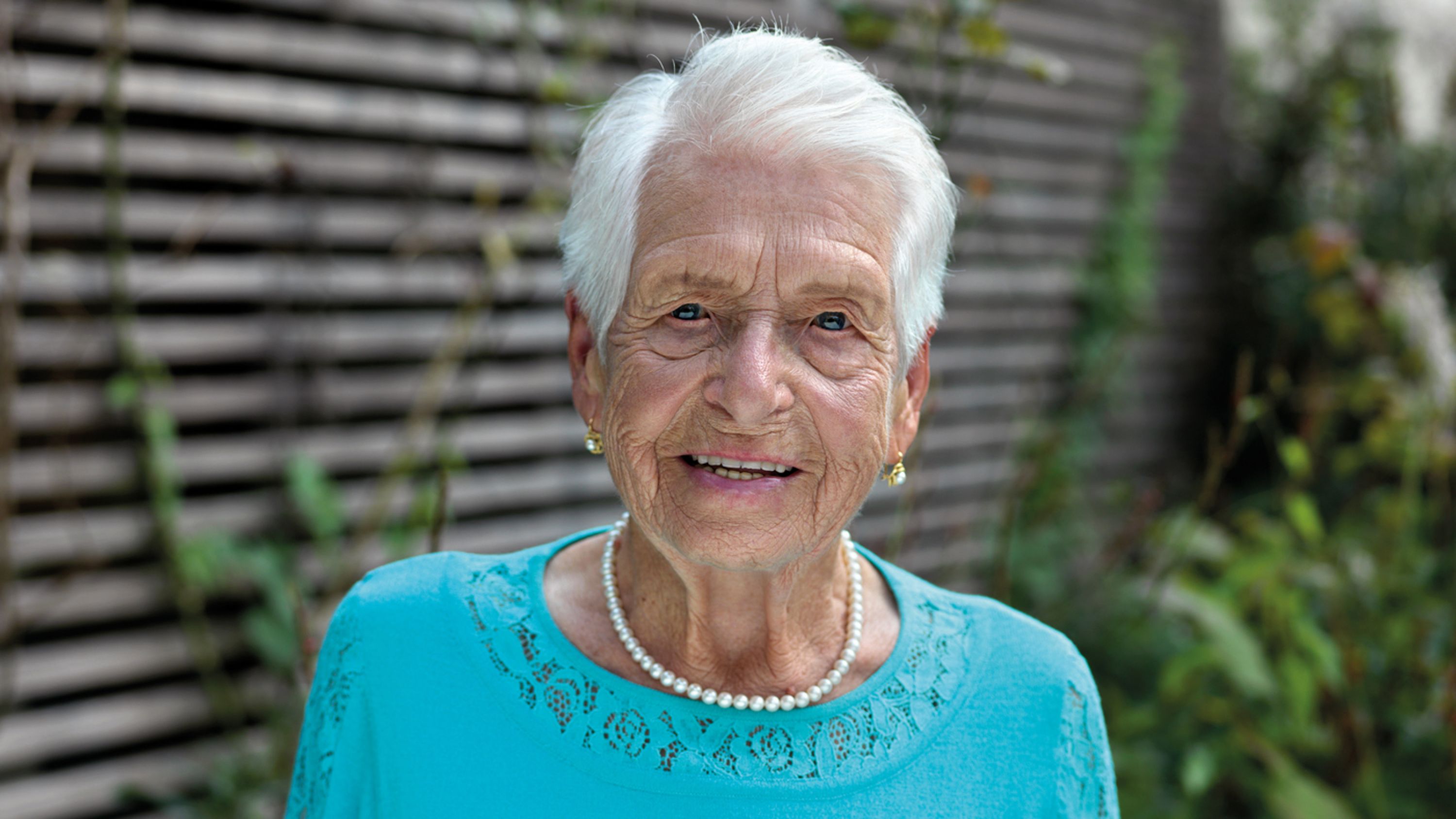 Eine Seniorin mit blauem Oberteil und Perlenkette schaut lächelnd in die Kamera.